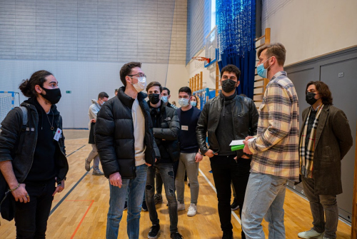 Studenci stoją i rozmawiają z wykładowcą na hali sportowej.