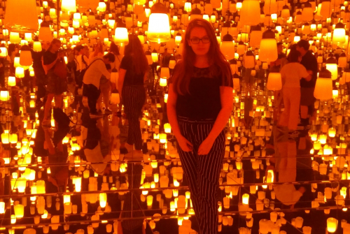 Studentka stoi. Nad nią wiszą świecące lampy, które odbijają się w lustrzanych ścianach.