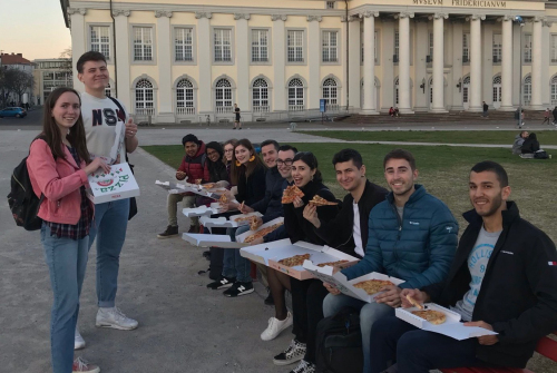Uśmiechnięci studenci trzymają pudełka z pizzą. Dwie osoby po lewej stroją. Pozostałe osoby po lewej siedzą.