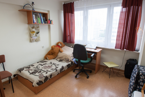 Wnętrze pokoju w akademiku. Po lewej stronie zdjęcia łóżko oraz biurko.