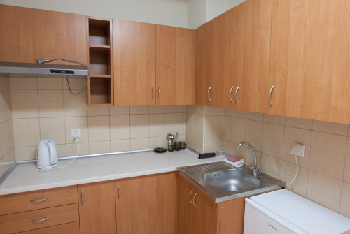 Wnętrze aneksu kuchennego. Pomieszczenie wyposażone w szafki dolne oraz wiszące, czajnik elektryczny, zlew, okap i lodówkę.