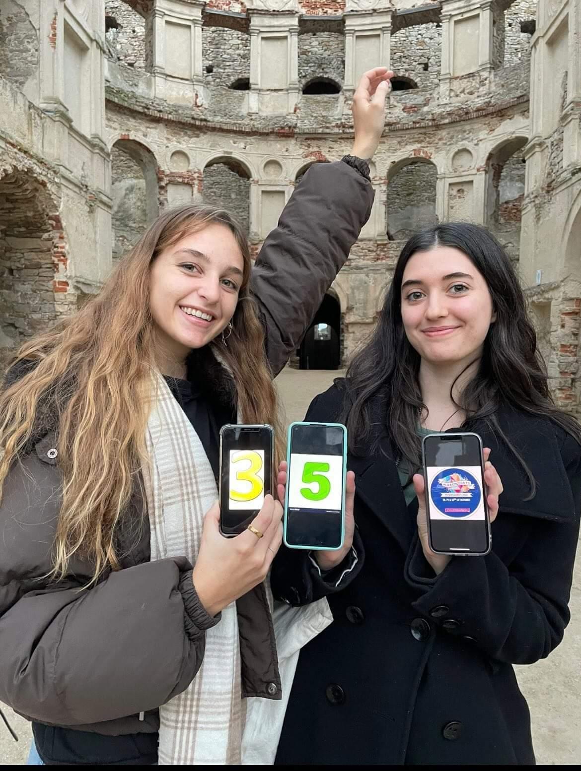 Dwie studentki trzymają telefony z wyświetlonym logo Erasmus Days. W tle ruiny zamku.