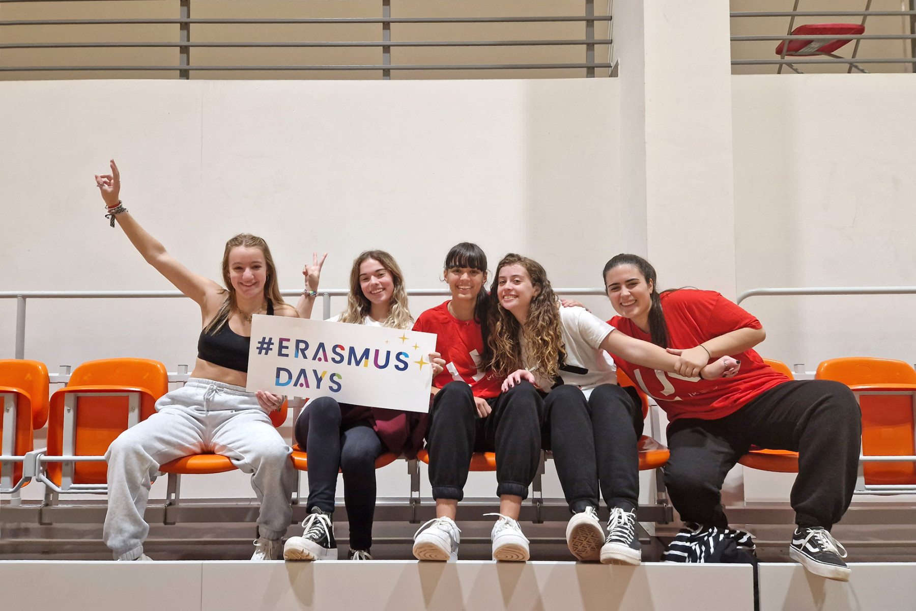 Grupa dziewcząt pozuje do zdjęcia siedząc na trybunach. Jedna z nich trzyma planszę na napisem Erasmus Days.