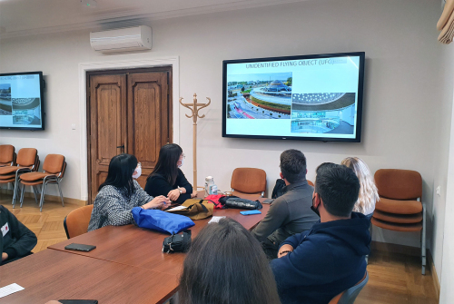 Studenci siedzą. W tle widać ekran telewizora. Studenci oglądają wyświetlaną na nim prezentację.