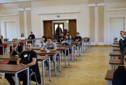 Studenci siedzą. Ławki ustawione są w rzędach. Zdjęcie z oddali.