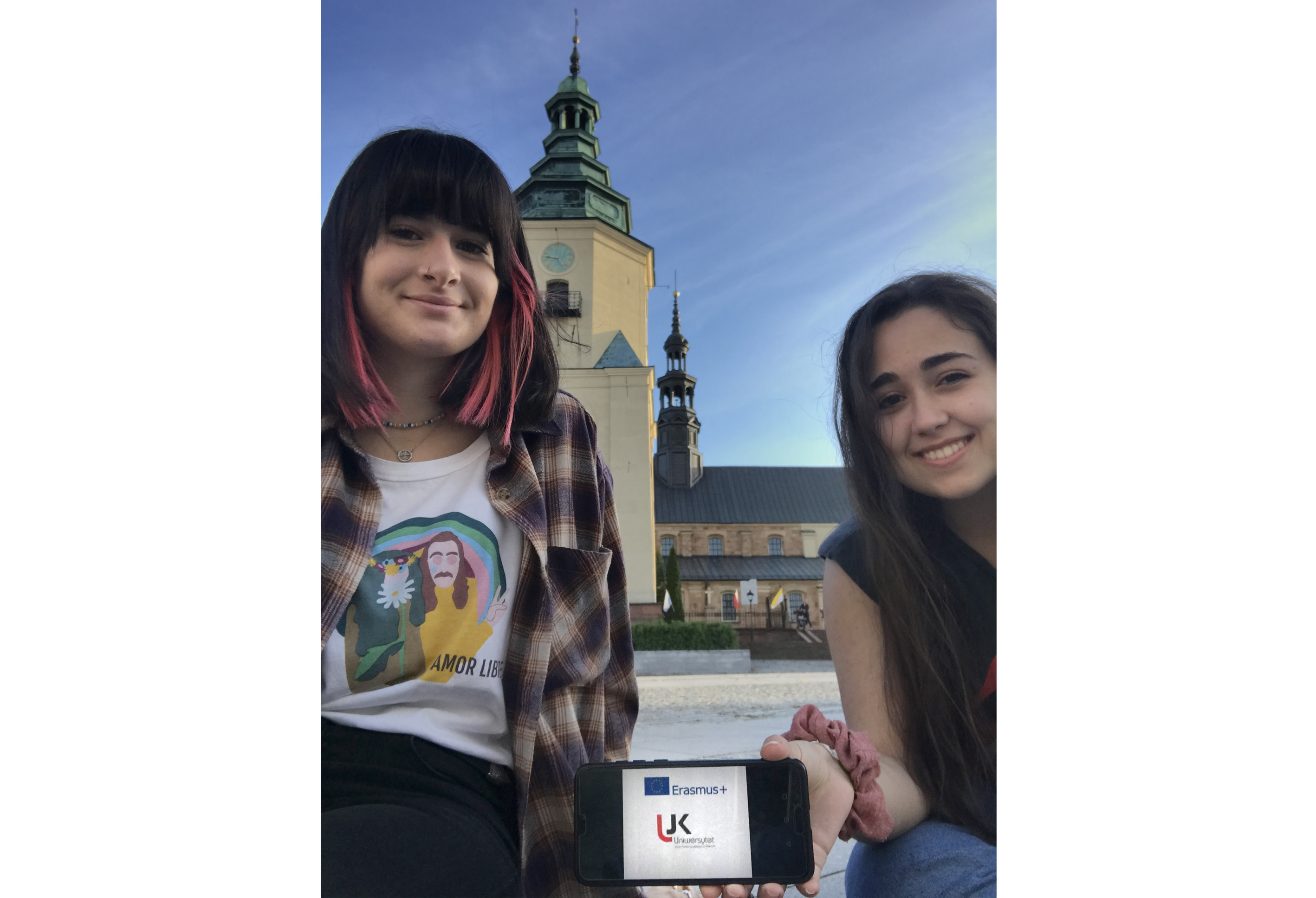 Dwie studentki siedzą. Trzymają telefon. W tle budynek Katedry.