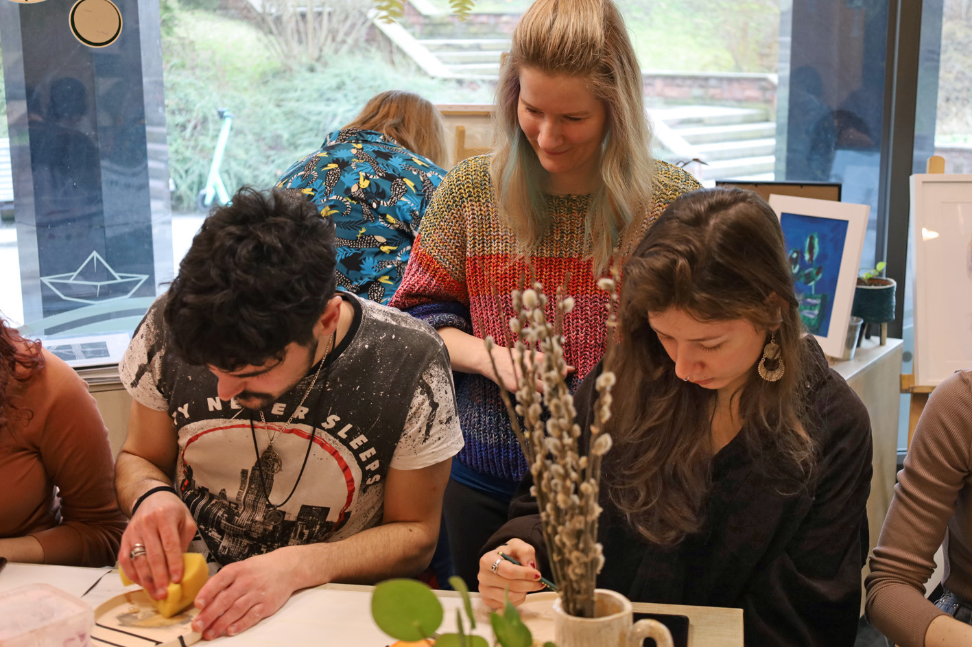 studenci siedzą wokół stołu warsztatowego i malują ceramiczne miseczki.