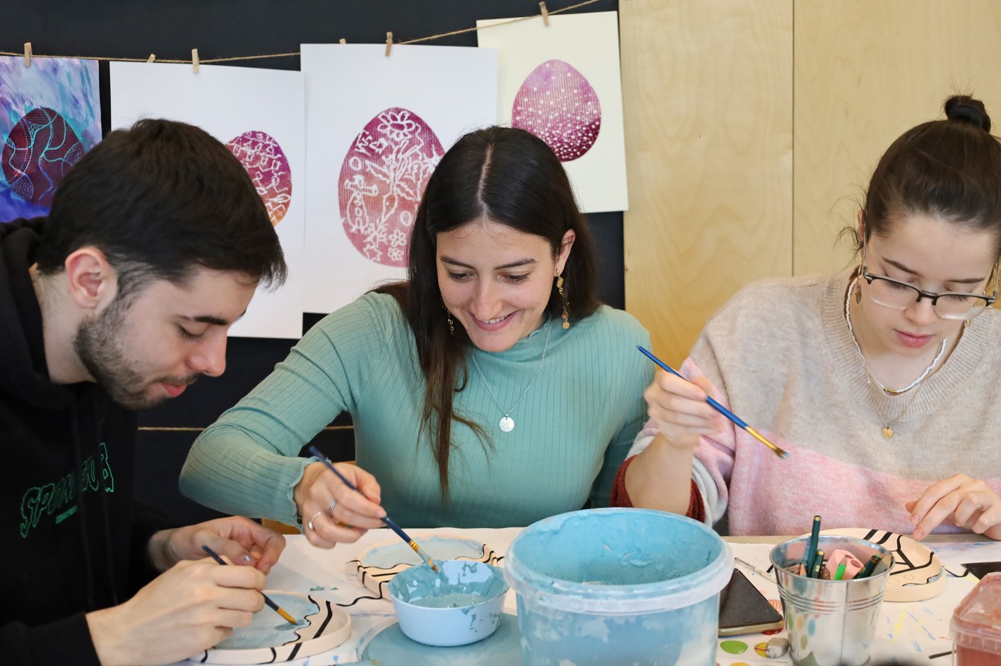 studenci siedzą wokół stołu warsztatowego i malują ceramiczne miseczki.