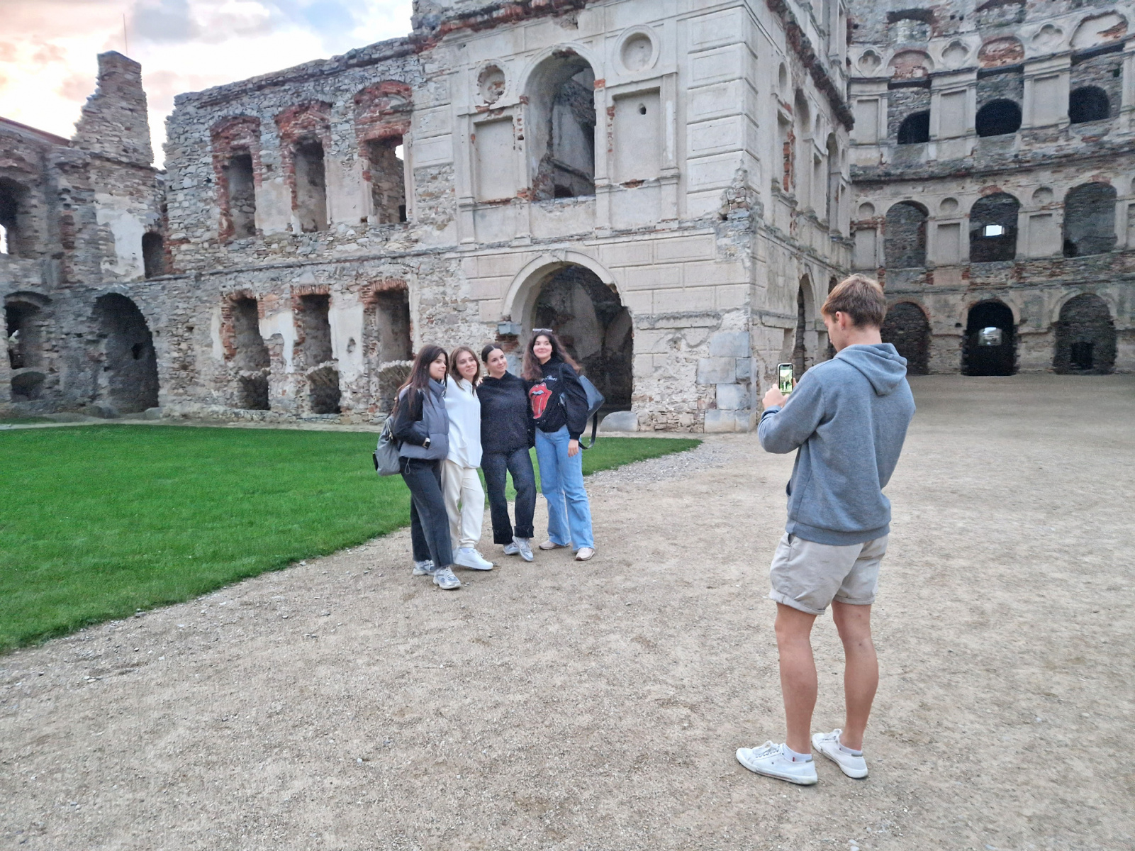 Po lewej grupa studentek pozuje do zdjęcia. Po prawej student stoi z telefonem i robi im zdjęcia. W tle ruiny zamku Krzyżtopór.